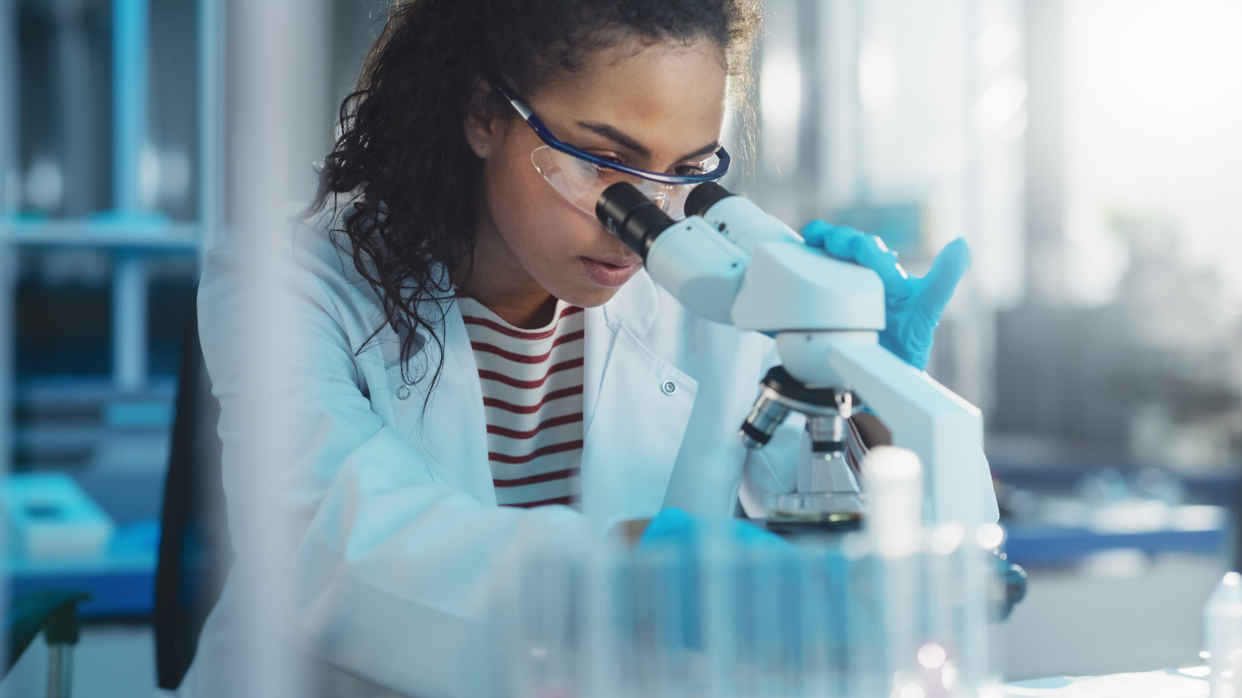 Scientist in a lab coat and goggles examines a sample under a microscope, highlighting the focus on research and discovery in a laboratory setting.